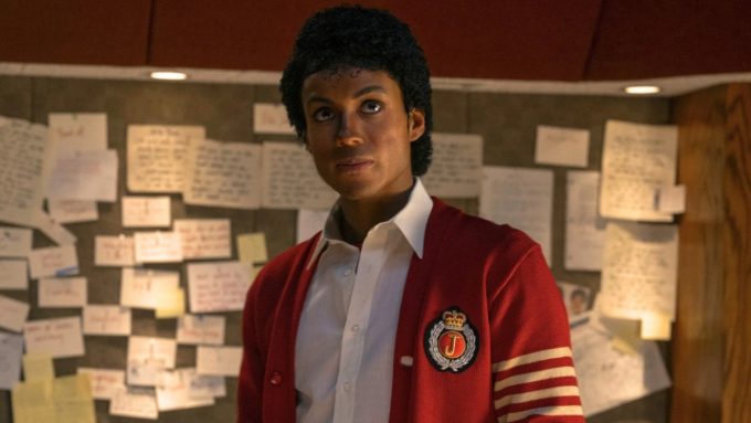 A production still featuring Jaafar Jackson portraying Michael Jackson. He is standing in a room with a wall covered in handwritten notes and papers. He wears a red cardigan with a "J" crest and white stripes on the sleeve over a white collared shirt, sporting a signature curly hairstyle.