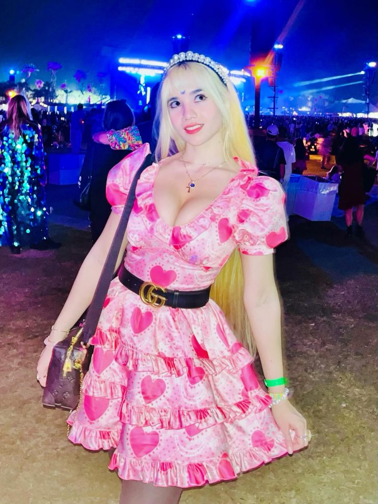 Windy Girk at an outdoor night event with long blonde hair and a beaded headband. She is wearing a pink ruffled dress with a heart pattern, cinched with a black belt featuring a gold "G" buckle.