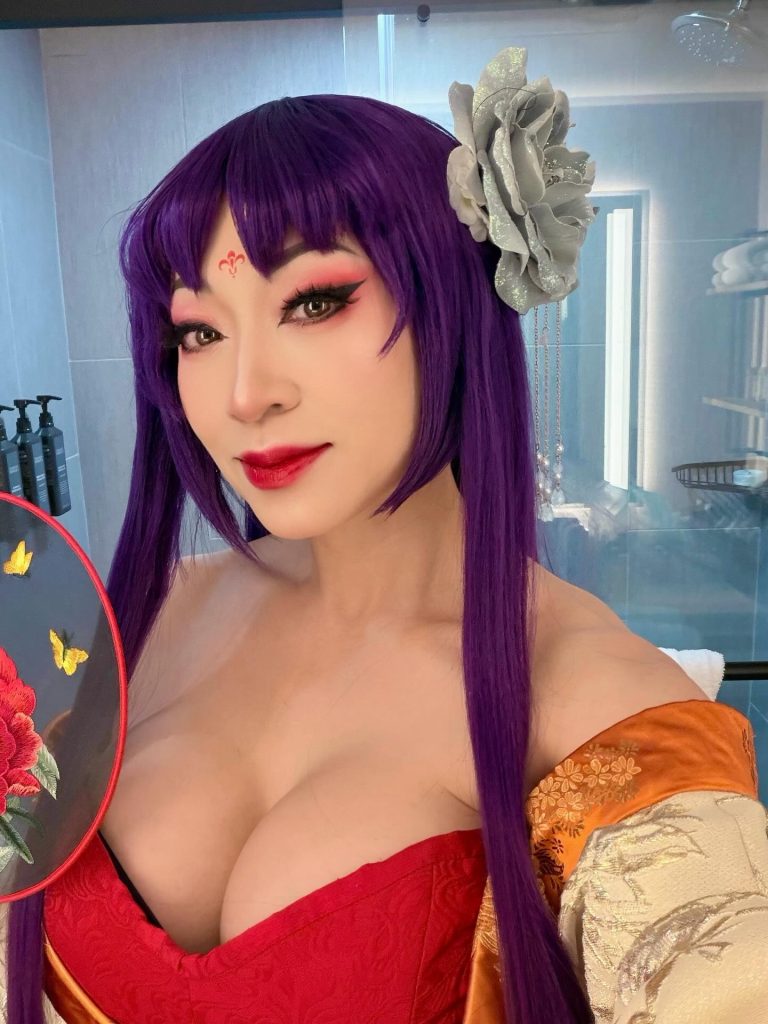 A close-up selfie of Yaya Han sporting a purple wig with straight bangs and a large silver floral accessory. She wears a red and gold patterned kimono-style top with striking red eye makeup and a small red symbol on her forehead. She holds a traditional decorative fan, posing in a brightly lit room.