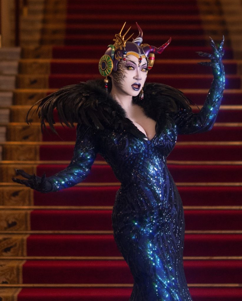 An atmospheric shot of Yaya Han in a masterful recreation of Edea Kramer from Final Fantasy VIII. Standing on a red-carpeted grand staircase, she wears a shimmering, form-fitting navy blue sequined gown with a dramatic black feathered collar. Her elaborate headpiece and intricate facial prosthetics showcase the high-level craftsmanship that defines her work.