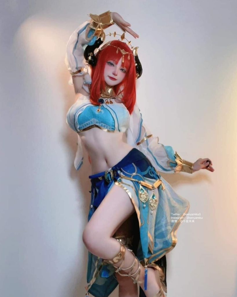 A full-body shot of Senyamiku cosplaying as Nilou from Genshin Impact. She has long red hair and is wearing an intricate blue and white dancer's costume with gold jewelry, horns, and a translucent veil. She is posing with one hand above her head against a plain white wall.