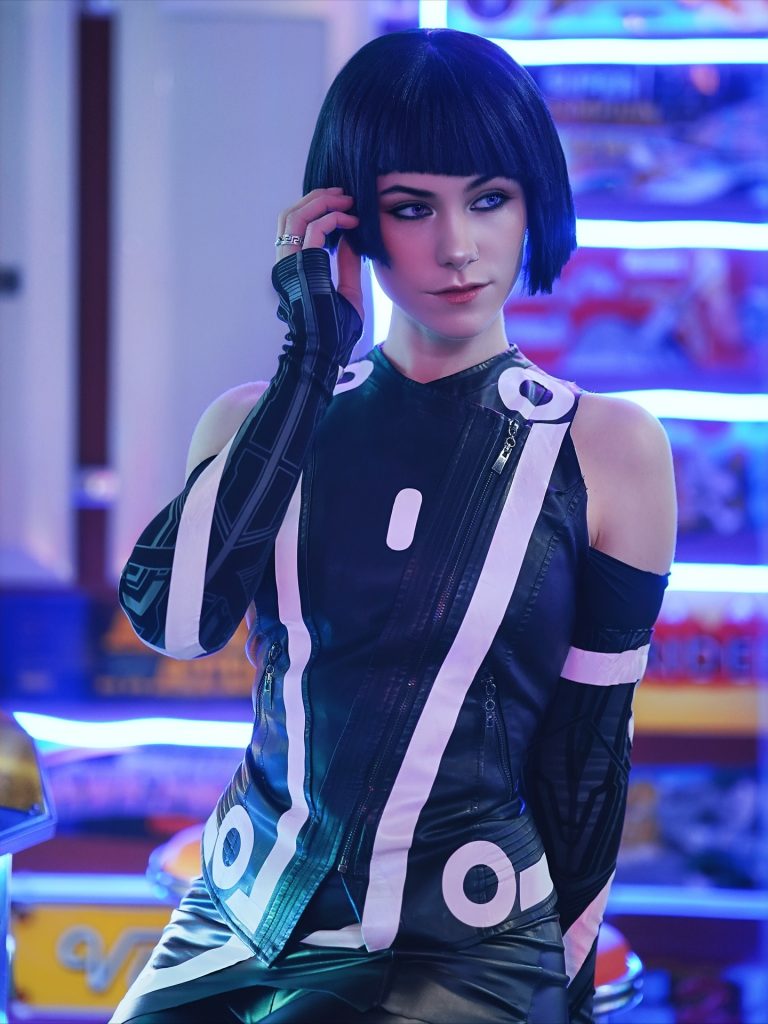 A woman with a short black bob and bangs wearing a black and white futuristic, sleeveless high-neck top. She has purple contact lenses and is posing against a blurred background of blue neon lights.