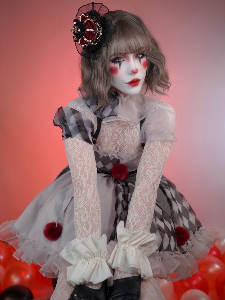 A woman in clown-inspired makeup and costume. She has white face paint with red accents, a grey wavy bob, and is wearing a ruffled black and white checkered dress with red pom-poms.