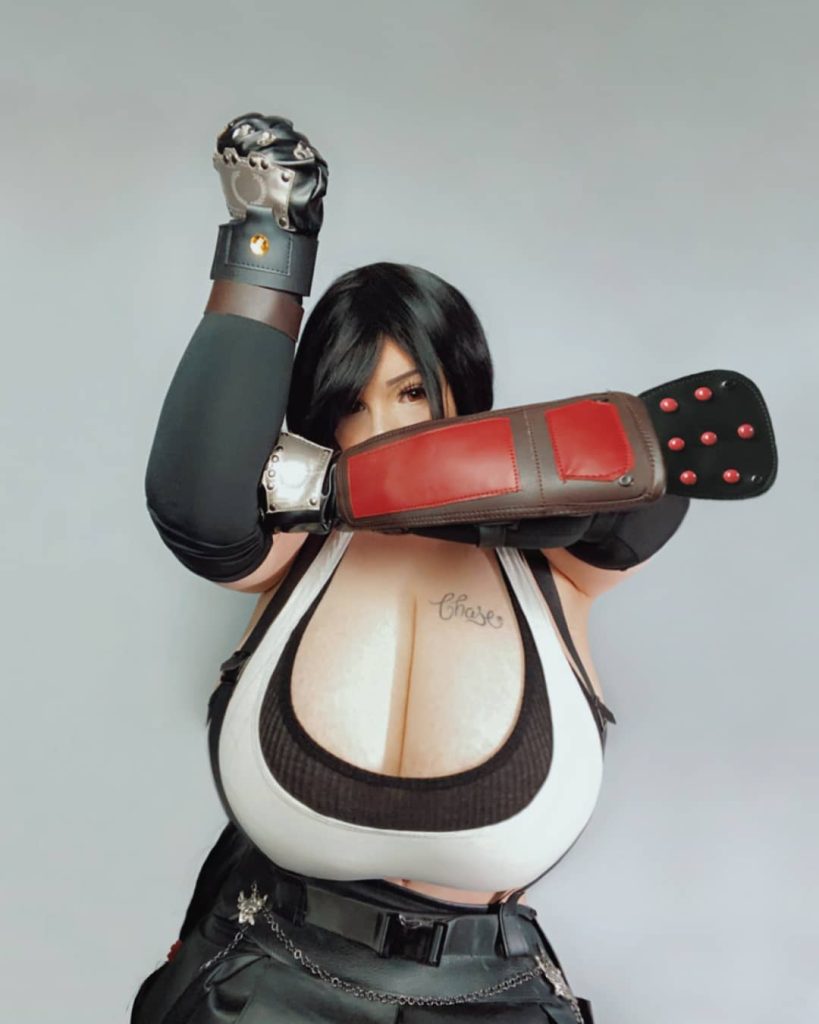 A woman in a Tifa Lockhart cosplay from Final Fantasy VII. She is wearing a white tank top, black suspenders, and armored gauntlets, posing with one arm raised and the other across her face.