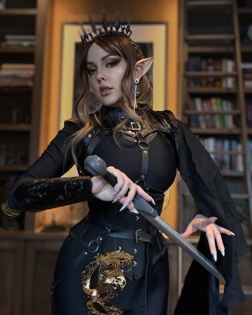 A woman dressed in a dark, regal elf cosplay. She is wearing a black high-collared outfit with a leather harness, a crown of dark spikes, and pointed elf ears while holding a sword.