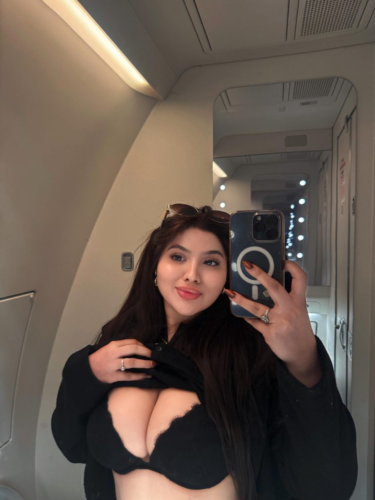An Asian OnlyFans influencer mirror selfie taken inside what appears to be a plane cabin, showing Dea Ayu smiling while pulling up her black shirt to reveal her bra.