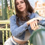 A casual outdoor portrait of popular cosplayer Enji Night leaning against a green bridge railing while looking into the camera. She wears a cropped denim jacket over a white top and light-wash jeans, capturing the essence of her lifestyle content often teased on platforms like Enji Night OnlyFans.