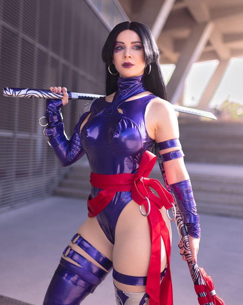 A striking studio portrait of Enji Night cosplaying Psylocke from X-Men, wearing a glossy purple bodysuit and holding a katana over her shoulders. This exclusive look highlights the bold comic book content shared with subscribers on her Enji Night OnlyFans.