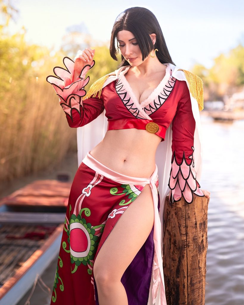An elegant outdoor photoshoot of Enji Night cosplaying Boa Hancock from One Piece. Posing near a wooden dock and water, her detailed red and pink costume captures the character's signature look