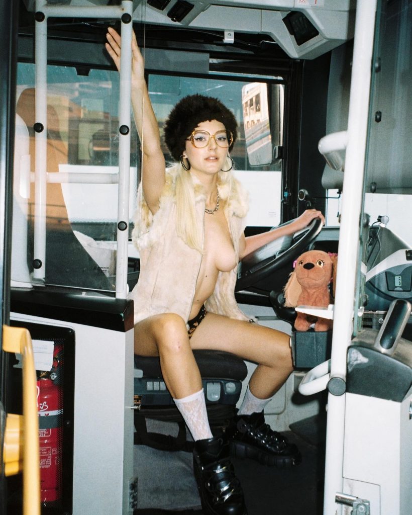 Stylized creative photoshoot of popular internet celebrity Eva Elfie OnlyFans star sitting in a driver's seat wearing a fur hat, glasses, and an open vest.