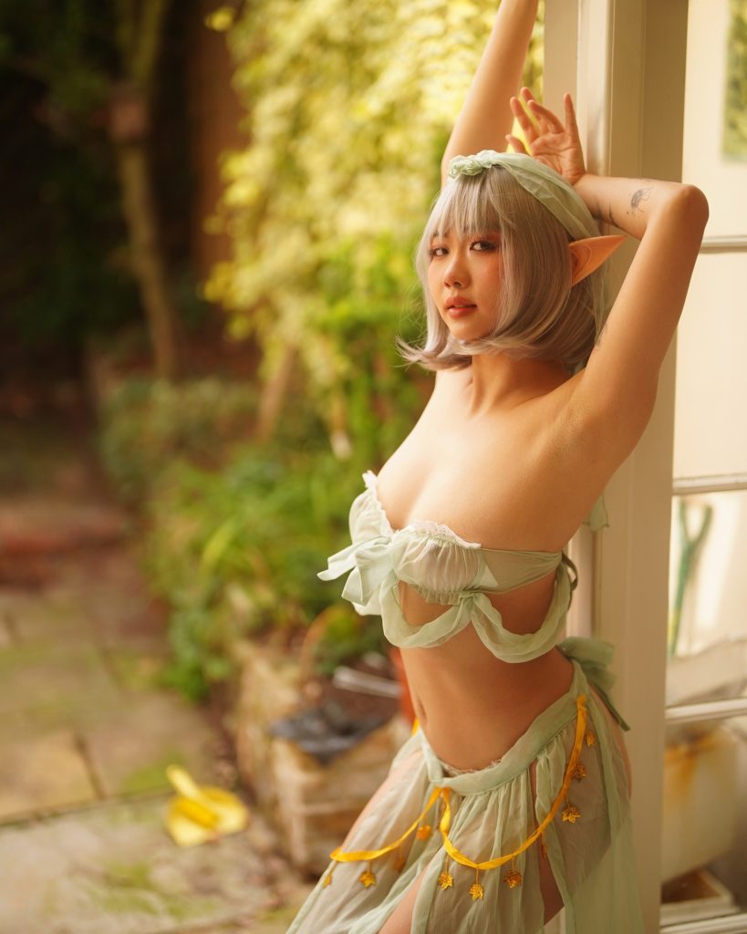 An asian onlyfans model posing in an outdoor setting while wearing a light green elven cosplay outfit. The ensemble includes pointed elf ears, a sheer ruffled top, and a matching skirt with gold leaf accents.