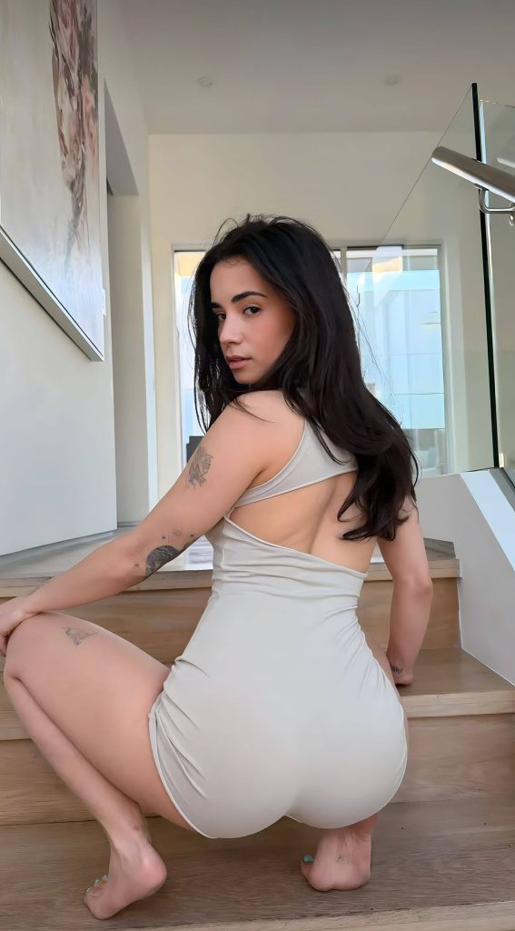 In this striking image, model Jameliz Benitez Smith, whose exclusive photo shoots on OnlyFans have a massive following, crouches on a modern wooden staircase, turning to look back. She wears a light beige cut-out bodysuit and showcases her multi-tattooed arms.