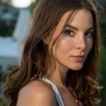 A close-up headshot of Kacey Clarke looking directly at the camera with a subtle smile. Her long, wavy brown hair frames her face, highlighted by soft, natural outdoor lighting.