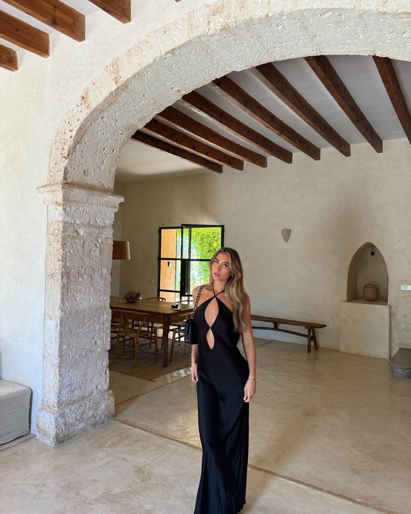 Lily Phillips standing under a large stone archway inside a rustic, Mediterranean-style room with exposed wooden ceiling beams and a large dining table in the background. She is wearing a long black halter dress with a keyhole cutout and carrying a black shoulder bag, capturing a moment from the Lily Phillips OnlyFans journey from Instagram to fame.