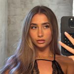 A close-up mirror selfie of Lily Phillips looking off to the side while holding a black smartphone. She is wearing a delicate black lace halter top and a gold clover necklace, with her long blonde-highlighted hair falling over her shoulder. This image highlights a candid moment from Lily Phillips' OnlyFans and social media presence.