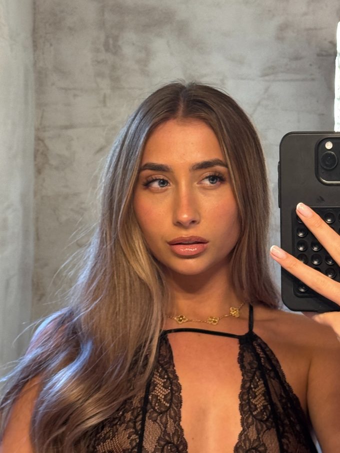 A close-up mirror selfie of Lily Phillips looking off to the side while holding a black smartphone. She is wearing a delicate black lace halter top and a gold clover necklace, with her long blonde-highlighted hair falling over her shoulder. This image highlights a candid moment from Lily Phillips' OnlyFans and social media presence.