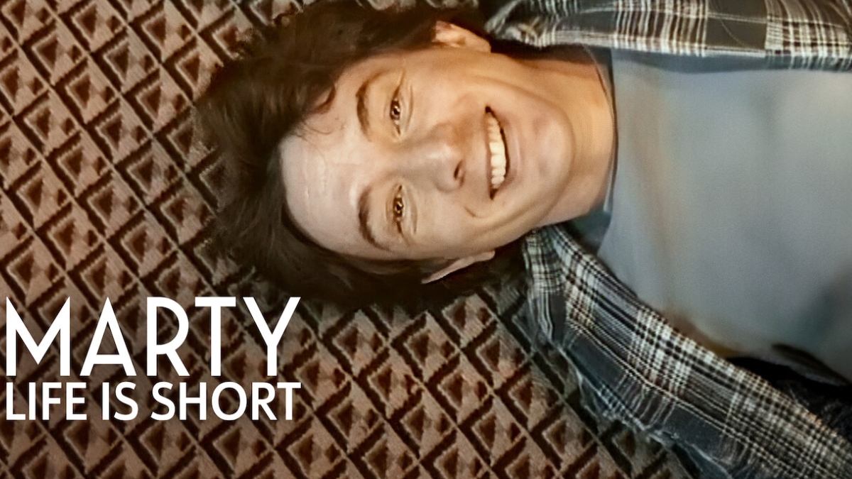 A smiling young Martin Short lying on a patterned carpet while wearing a plaid shirt, featuring the overlaid text "MARTY LIFE IS SHORT."