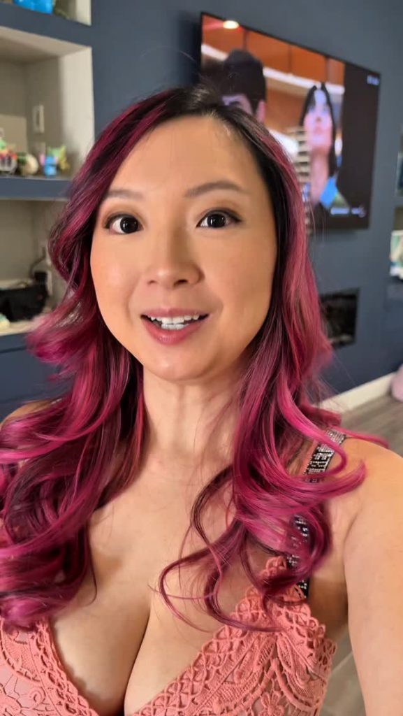 A close-up selfie of Monique Mae featuring her vibrant pink and purple hair, smiling while wearing a peach-colored lace top in a modern home setting.