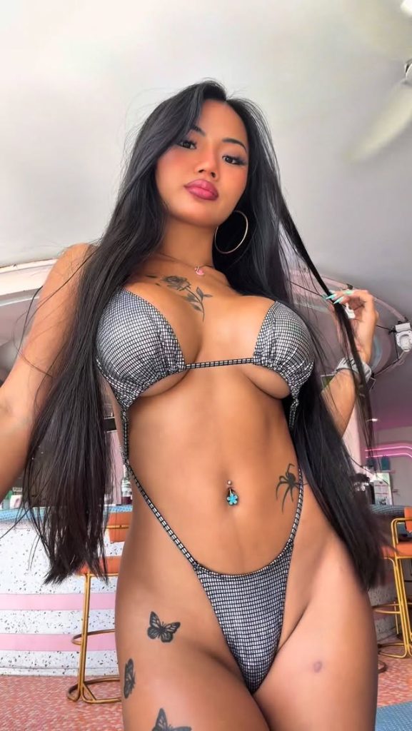 A striking low-angle portrait of Praew Asia OnlyFans looking confidently at the camera. She is wearing a metallic grey, grid-patterned micro bikini that highlights her spider and butterfly tattoos.