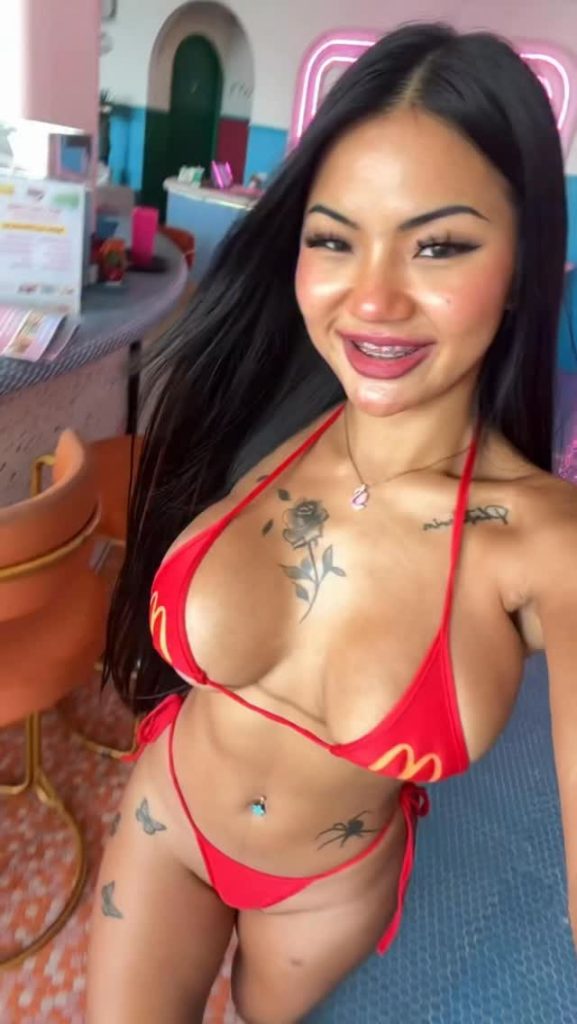 A cheerful selfie of Praew Asia OnlyFans showing off a vibrant red string bikini featuring the iconic yellow McDonald's arches. Her distinctive rose tattoo on her chest and butterfly hip tattoos are on full display.