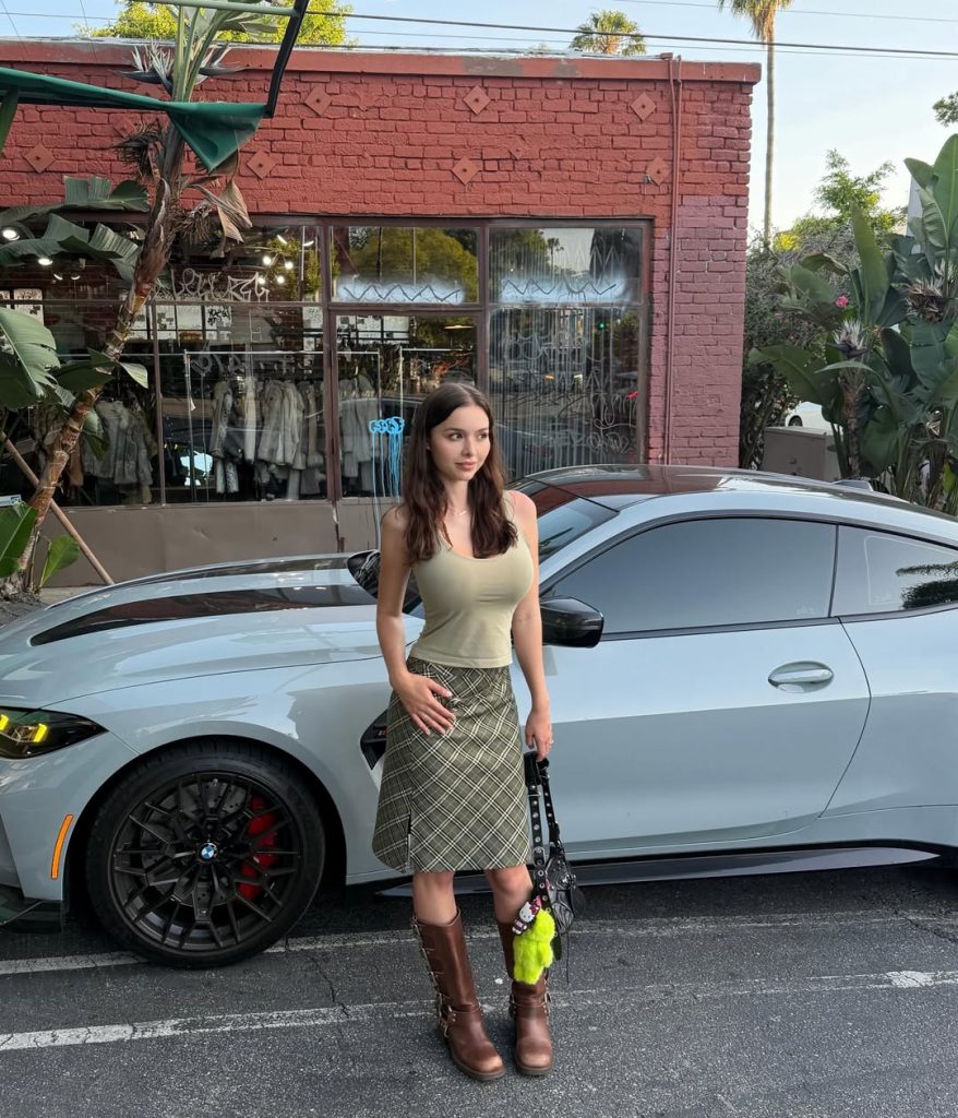 A casual street style photo of Sophie Mudd OnlyFans standing outdoors in front of a sleek grey sports car. She is wearing a green tank top, a plaid patterned skirt, and brown leather boots while holding a black handbag.