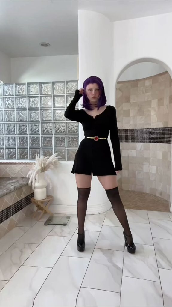 A full-body shot of Cosplay Influencers featuring Sunny Ray in a minimalist Raven cosplay from Teen Titans, wearing a black long-sleeved romper, purple wig, and sheer black stockings.