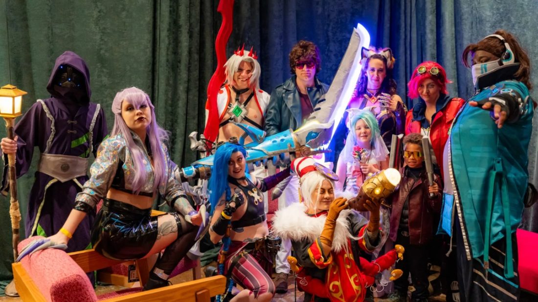 A diverse group of people posing in various intricate cosplay costume designs, featuring colorful wigs, elaborate props like oversized glowing swords, and themed outfits against a dark curtain backdrop.