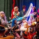 A diverse group of people posing in various intricate cosplay costume designs, featuring colorful wigs, elaborate props like oversized glowing swords, and themed outfits against a dark curtain backdrop.