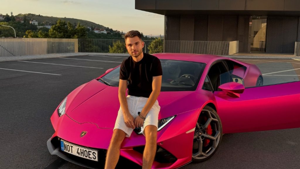 Adam Kajumi in a black t-shirt and white shorts sitting on the hood of a vibrant pink Lamborghini sports car in an outdoor parking lot, linked to a probe involving his OnlyFans Management Agency.