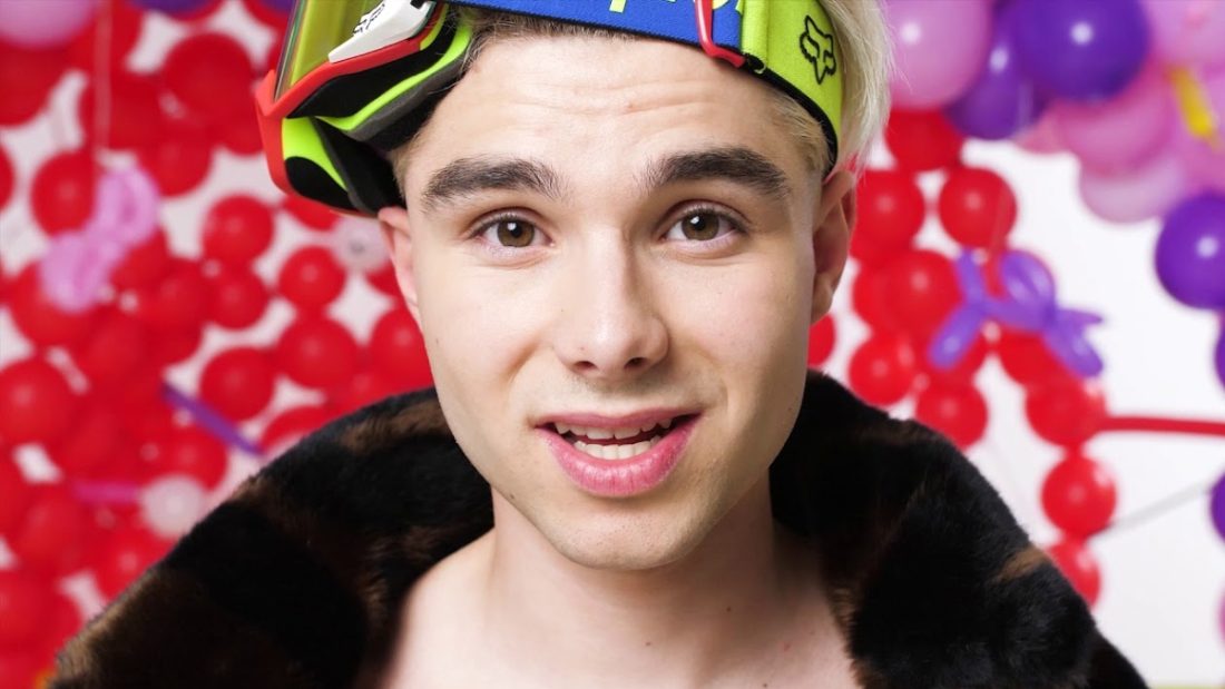 Close-up portrait of influencer Adam Kajumi wearing neon ski goggles and a fur-collared jacket against a background of red and purple balloons, related to investigations into his OnlyFans Management Agency.