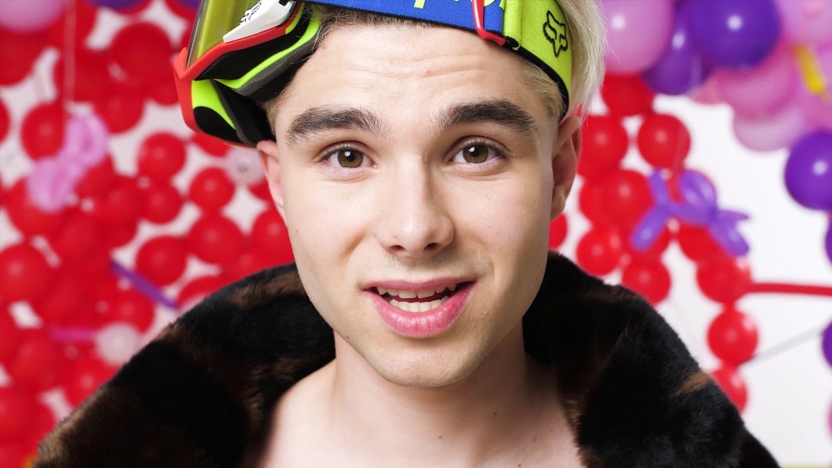 Close-up portrait of influencer Adam Kajumi wearing neon ski goggles and a fur-collared jacket against a background of red and purple balloons, related to investigations into his OnlyFans Management Agency.