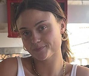 A close-up, slightly grainy headshot of a young woman with brown hair parted down the middle and pulled back. She is looking directly at the camera with a subtle, neutral smile. She has a nose septum ring piercing, a small stud piercing on her nostril, and wears multiple layered chunky gold hoop earrings. A gold chain necklace is visible around her neck, and she wears a white tank top. The background is softly blurred with a hint of a red frame structure behind her.
