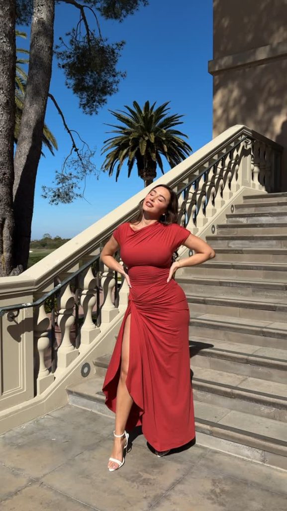 Ana Cheri OnlyFans content creator posing on stone outdoor stairs under a clear blue sky, wearing an elegant, form-fitting red gown with a high thigh slit.