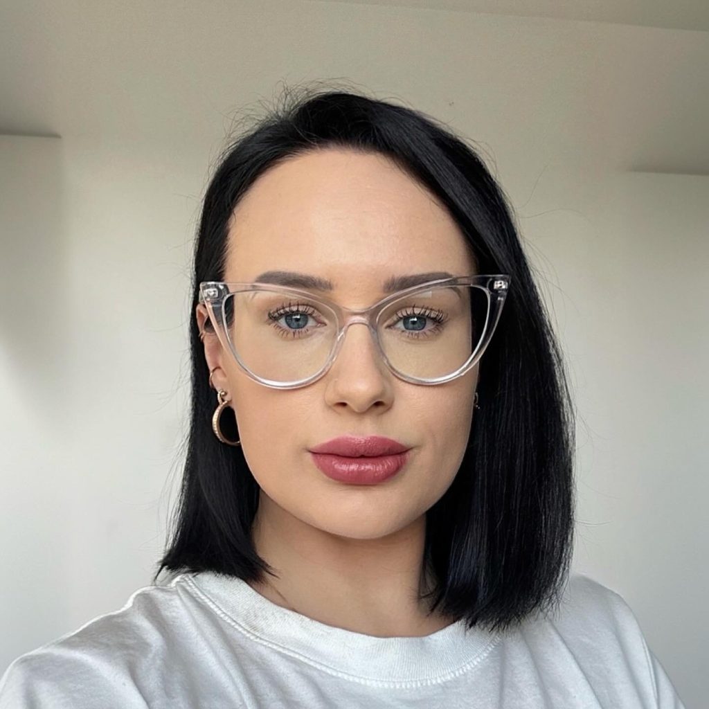 A close-up portrait of content creator Angel Wicky featuring dark hair, wearing clear-framed glasses, gold hoop earrings, and a white top against a plain background.