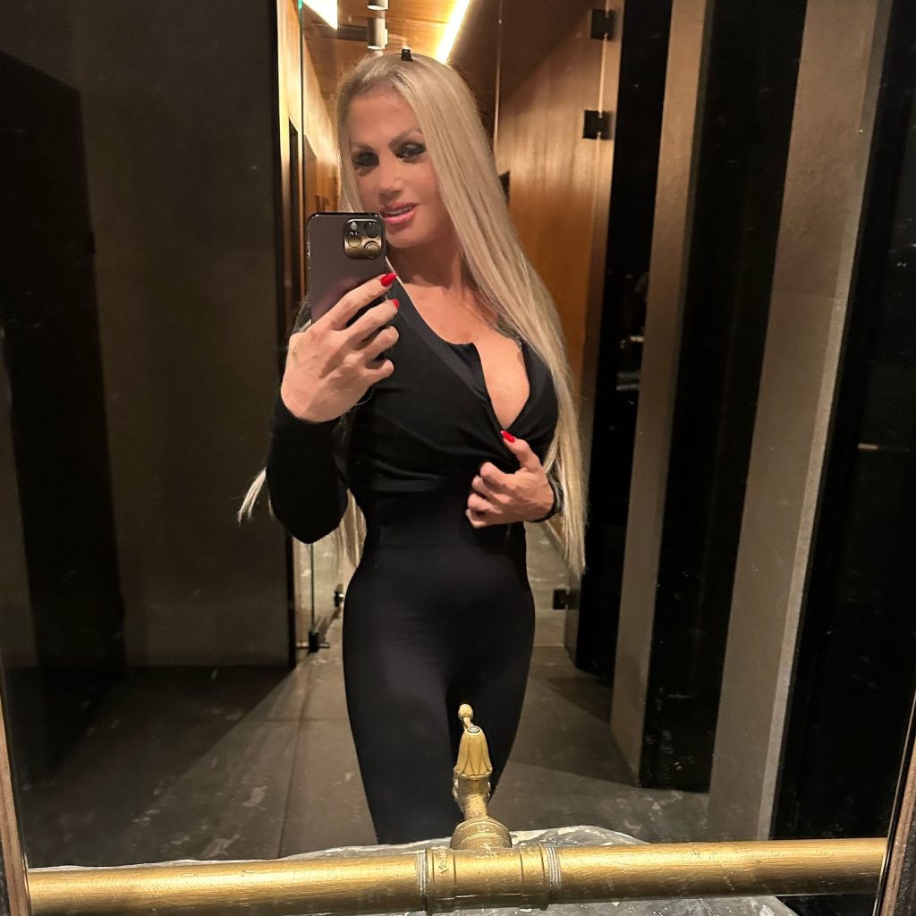 A mirror selfie of Angela Zisi, a well-known Greek Onlyfans creator, posing in a modern bathroom with wood paneling while wearing a tight, long-sleeved black jumpsuit unbuttoned at the chest.