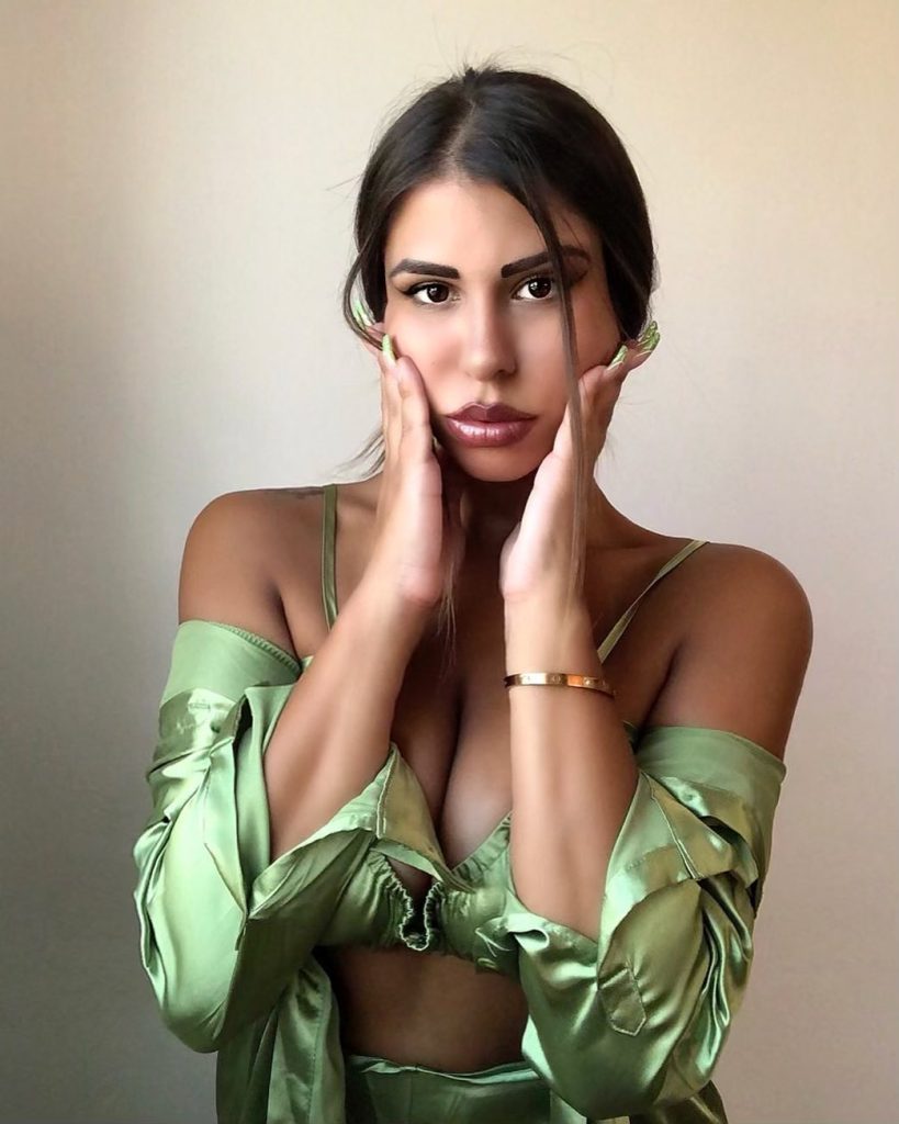 Greek Onlyfans model Angelina Piskopani looking into the camera while holding her face in a green satin crop top and matching robe.
