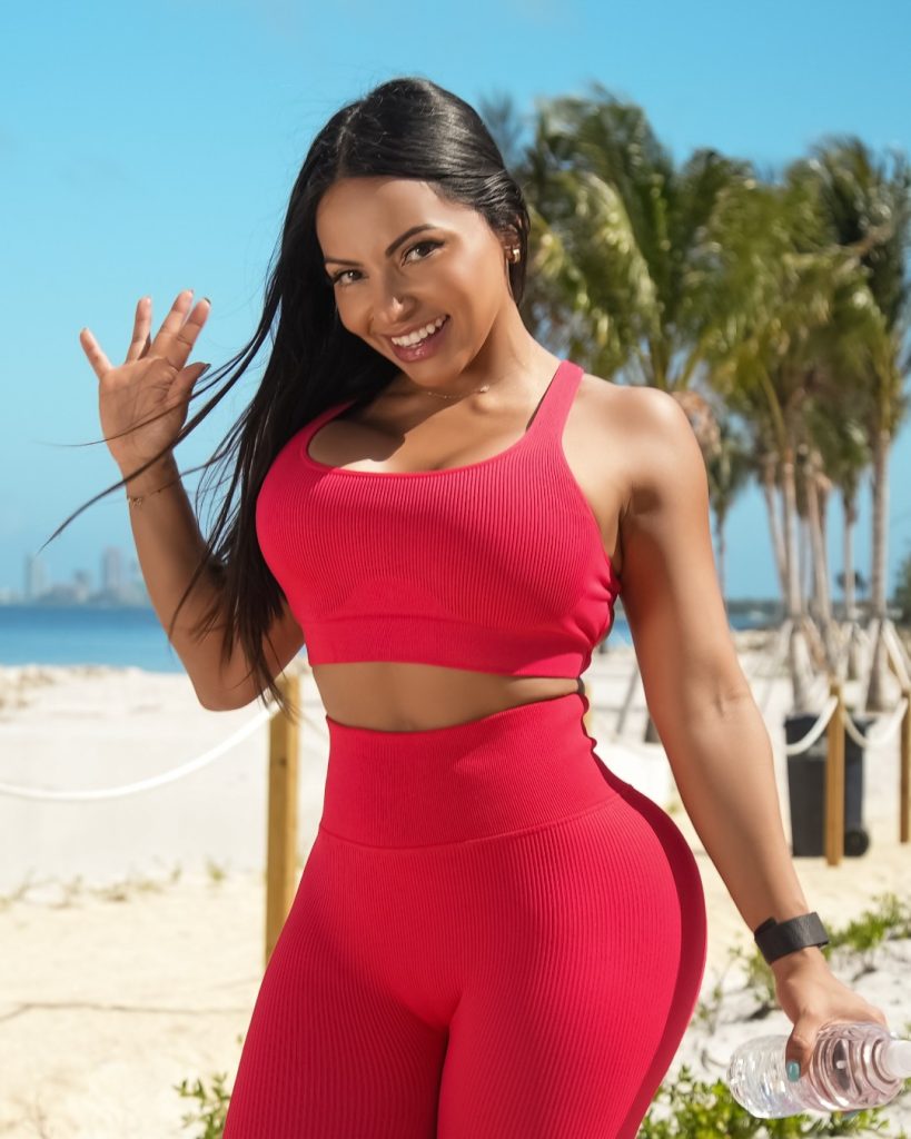 Brandy Salazar OnlyFans Model posing outdoors on a sunny beach, smiling and waving with palm trees and a blue sky behind her. She is wearing a bright red two-piece athletic crop top and leggings.
