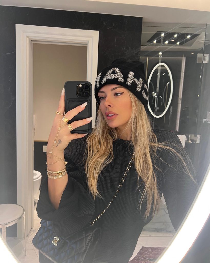 Mirror selfie of Corinna Kopf, popular content creator, wearing a black knit sweater and a black Chanel beanie with rhinestone lettering, looking aside while holding up her phone. A No PPV OnlyFans option for subscribers who want direct access.