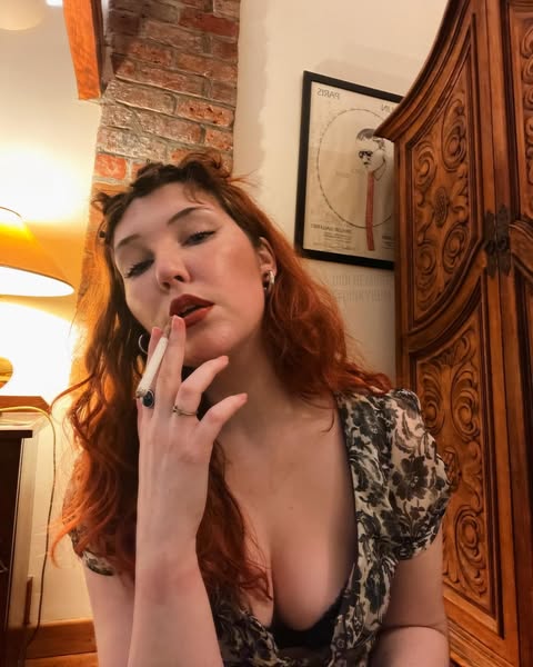 Close-up portrait of redheaded Irish OnlyFans model Didi Demure holding a cigarette to her lips while wearing a floral low-cut top next to a wooden wardrobe