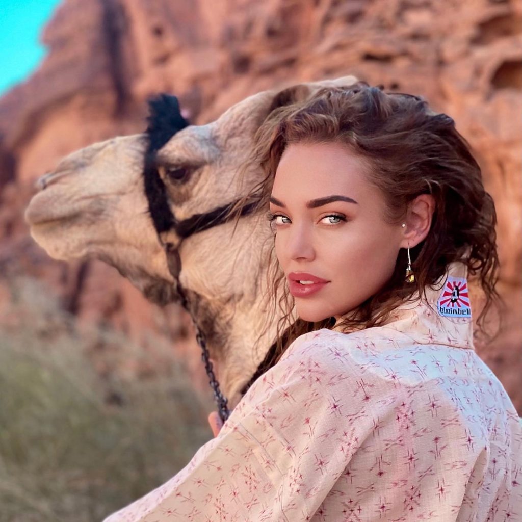 A portrait of Dorien Rose smiling while posing next to a camel in a desert landscape.