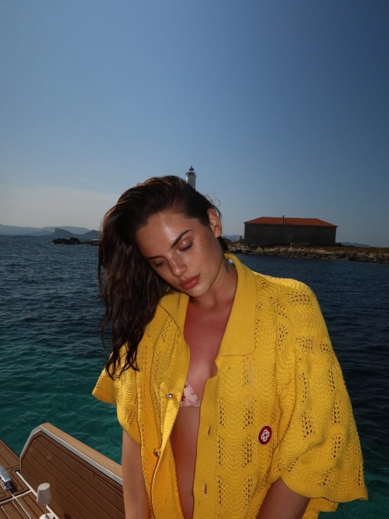 Famke Louise on a boat, wearing a yellow knit top over a patterned bikini, looking down with a lighthouse and small building visible on a rocky island in the background.