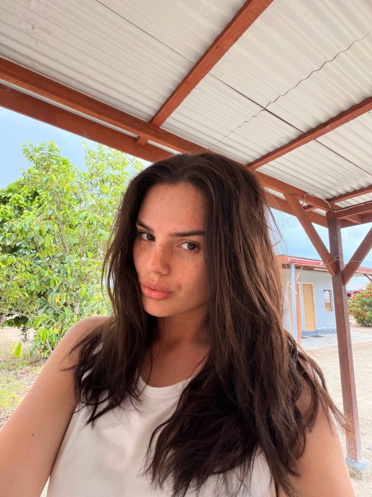 A selfie of Famke Louise with long dark hair, wearing a white tank top, posing under a wooden structure with trees in the background.