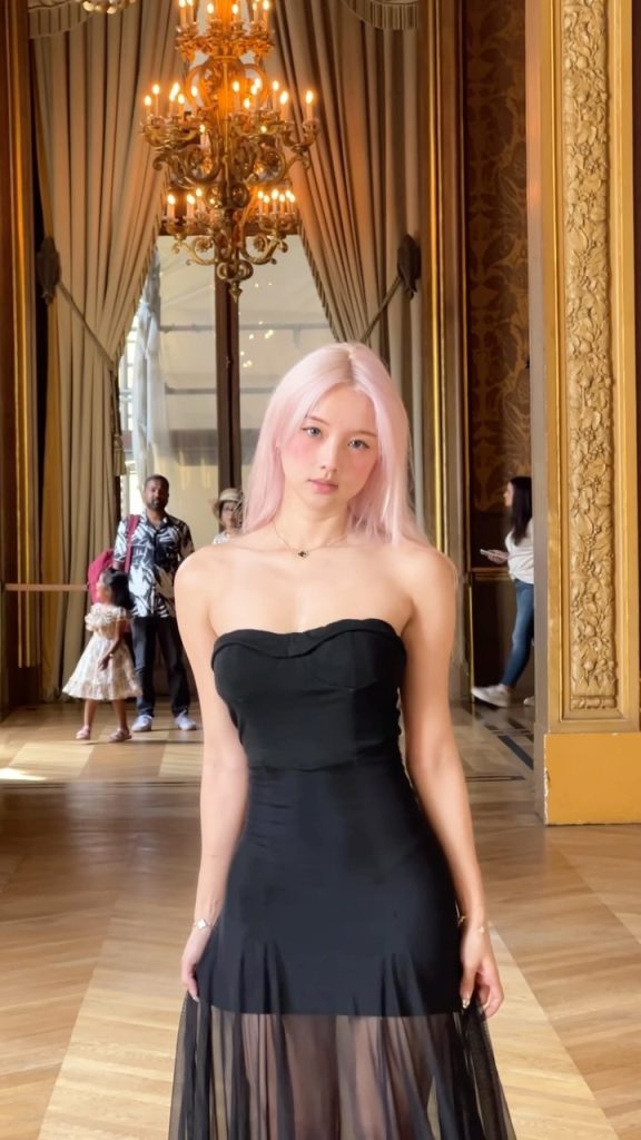 A photograph of a woman with pink hair looking straight at the camera, standing in an opulent hallway with a large, ornate crystal chandelier in the background. She is wearing a black strapless midi dress