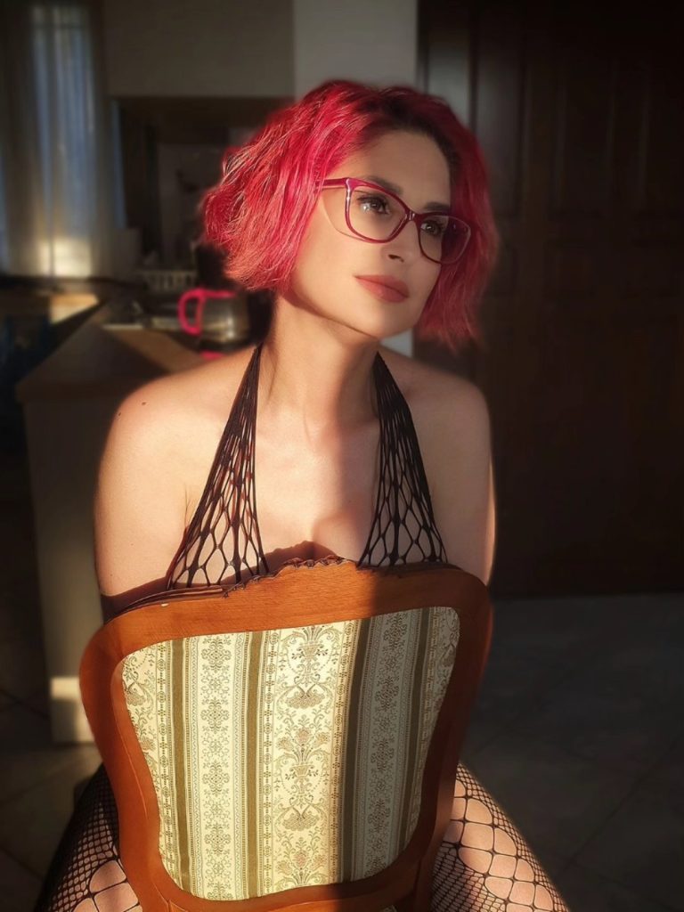 Elena Xatzi sitting backwards on an patterned vintage wooden chair under warm sunlight, wearing red glasses that match her short red hair and a black mesh halter top.
