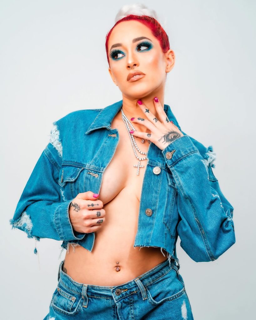 A studio shot of Elinnette OnlyFans Creator with vibrant red and white hair, wearing a distressed blue denim jacket open over her torso and matching denim jeans, accessorized with layered silver necklaces.