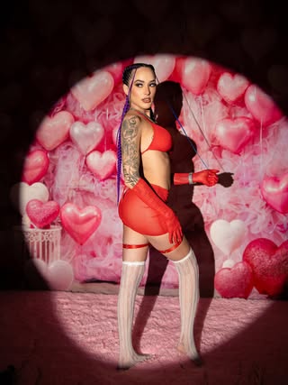 Elinnette OnlyFans Model posing in a red two-piece outfit with sheer gloves and white thigh-high stockings, illuminated by a spotlight against a backdrop of pink and red heart balloons.
