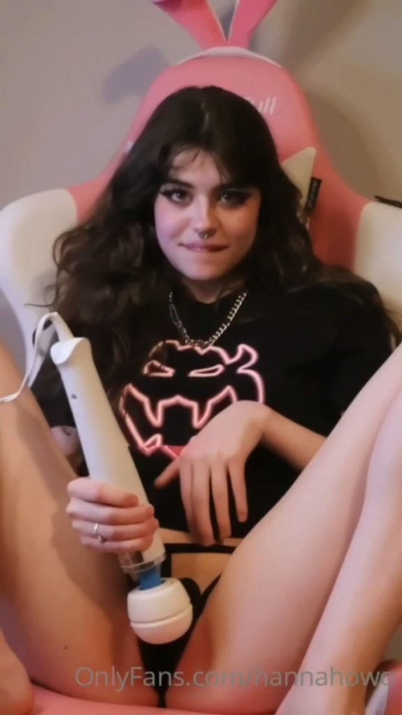 An image of content creator Hannah Kabel Onlyfans sitting in a pink and white gaming chair with bunny ears, wearing a black graphic t-shirt and holding a white handheld massage device.