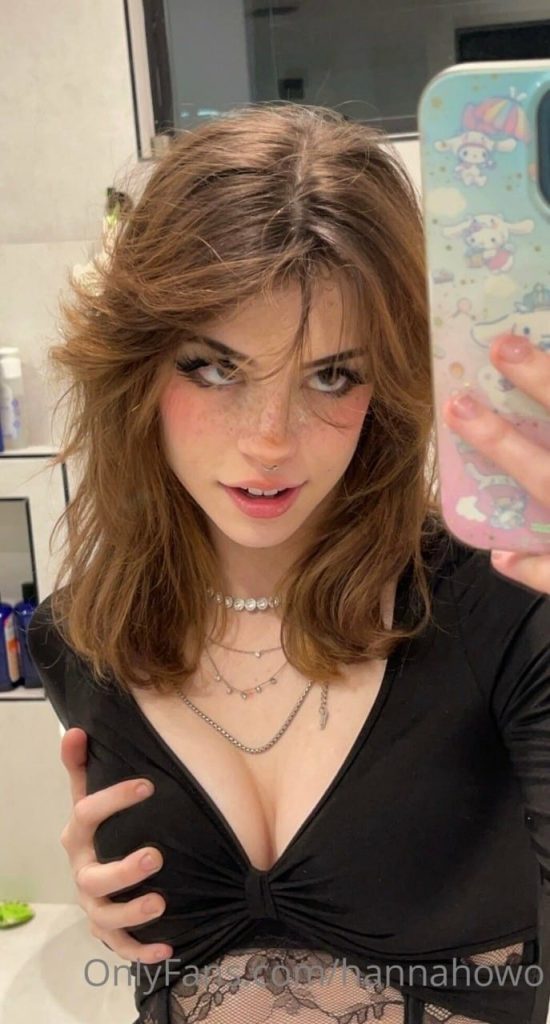 A close-up bathroom mirror selfie of Hannah Kabel Onlyfans with voluminous brown hair and freckles, wearing a low-cut black top and a silver chain necklace while looking into the camera.