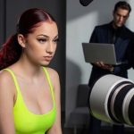 A young woman with dark red hair tied in a long ponytail sits under professional studio lighting during a media production shoot. She is wearing a bright neon green tank top and has a serious expression as she faces toward a large camera lens in the foreground. In the blurry background, two men in sharp business suits monitor the session—one looking at a laptop and the other holding a clipboard—illustrating the corporate management layer behind a highest earning OnlyFans creator.