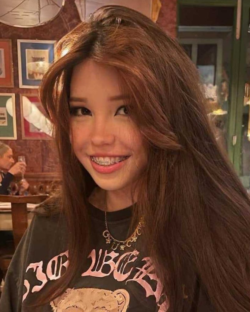 A candid, natural look at Internet personality Belle Delphine OnlyFans creator smiling in a restaurant, showing her natural brown hair, braces, and a casual black graphic t-shirt.